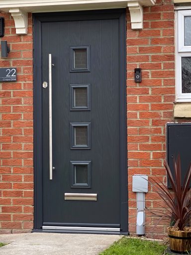 Dark grey front door with four small square windows and a vertical handle, set against brick.
